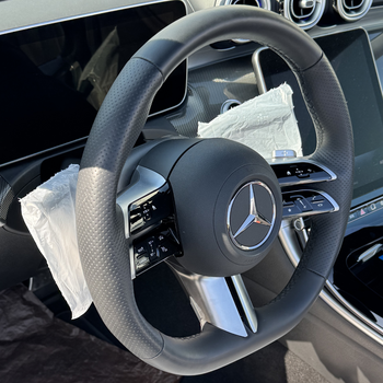 Das Bild zeigt das Lenkrad eines Mercedes-Benz im Fahrzeuginnenraum. Das Lenkrad ist schwarz mit silbernen Elementen und trägt in der Mitte das Mercedes-Logo. An den Speichen befinden sich Bedienelemente, und einige Teile sind noch mit dem Smartgrip3in1 Eco abgedeckt. Im Hintergrund sind Teile des Armaturenbretts und ein Display sichtbar.